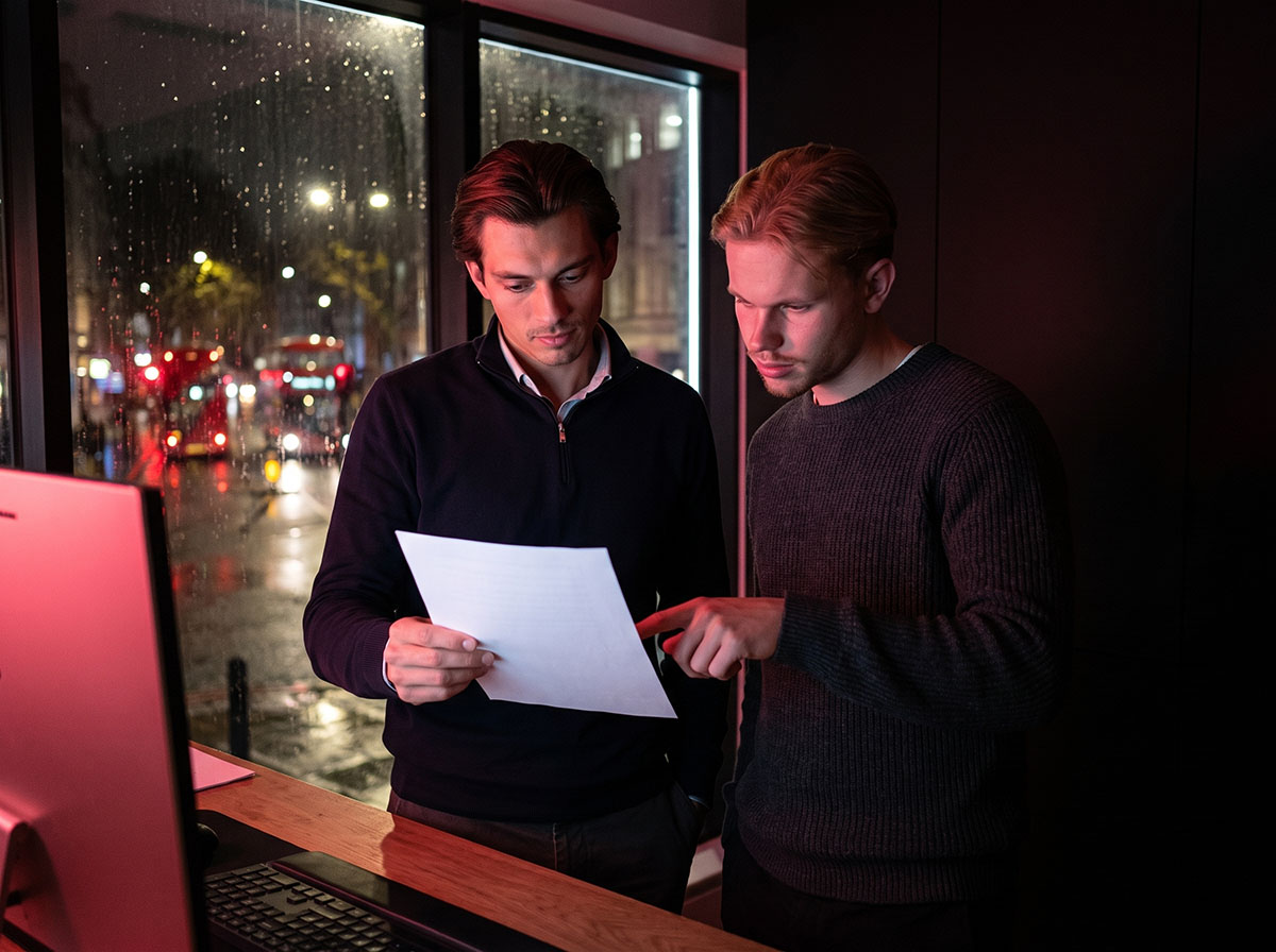 Two Forex Complex analysts reading the fine print of a prop firm rulebook by a rain-streaked London window